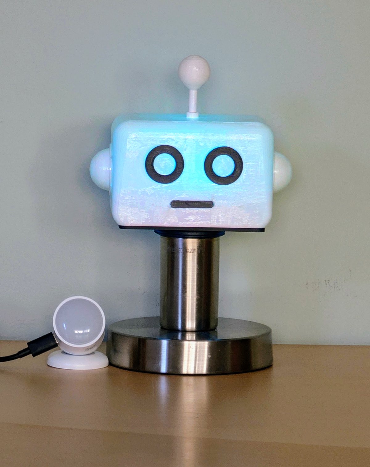 A translucent white 3D-printed robot head with two round dark eyes and a small antenna, lit faintly blue from inside, sitting on a steel bulb socket on a wooden sideboard, with a small round motion sensor beside it